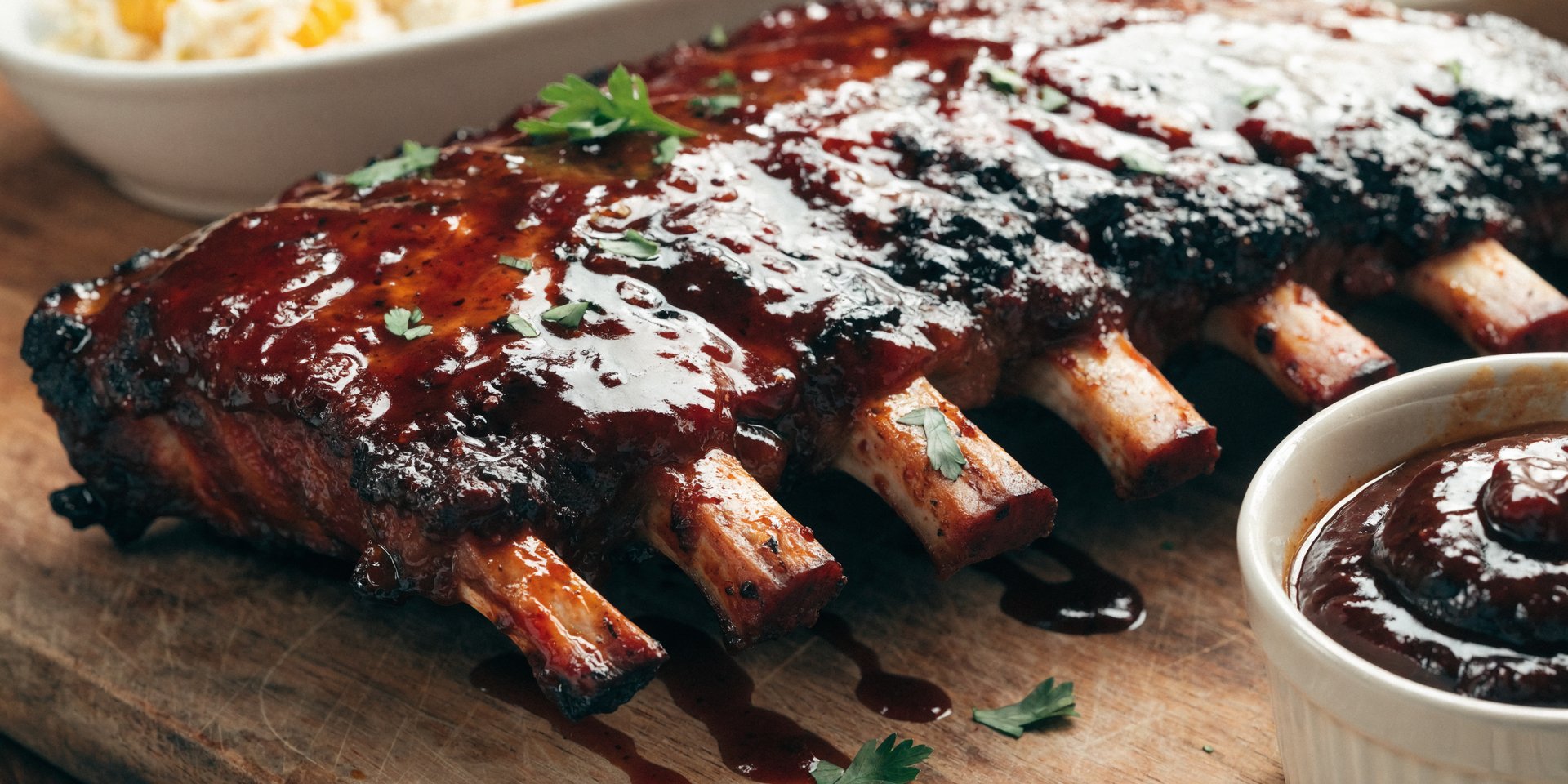 A rack of glistening, saucy BBQ pork ribs rests on a wooden cutting board, with a small dish of extra sauce nearby.